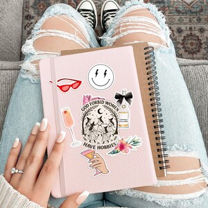 Puede incluir: Un cuaderno rosa adornado con pegatinas, incluyendo una carita sonriente, gafas de sol rojas y un gr&aacute;fico con el texto "GOD FORBID WOMEN HAVE HOBBIES". Un cuaderno marr&oacute;n con espiral descansa al lado. La persona lleva vaqueros azules rotos y zapatillas negras.