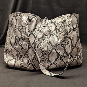 May include: A tote bag featuring a black and white snakeskin pattern. The bag has two shoulder straps and a drawstring closure at the top center. The material has a textured appearance with a repeating diamond-shaped scale design throughout.