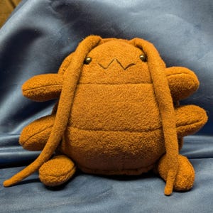 May include: A brown plush toy with a rounded body, four side limbs, and two long, dangling antennae. The toy features a stitched zigzag mouth and two small, dark, circular eyes. The fabric has a soft, textured appearance throughout the design.