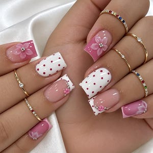 May include: Square-shaped nails featuring a mix of pink and white polish with red polka dots, three-dimensional pink flower accents with center crystals, and French tips. Several thin gold rings with small colorful stones are worn on the fingers.