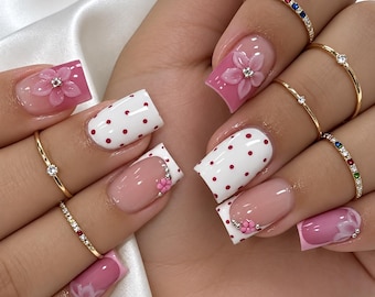 May include: Square-shaped nails featuring a mix of pink and white polish with red polka dots, three-dimensional pink flower accents with center crystals, and French tips. Several thin gold rings with small colorful stones are worn on the fingers.