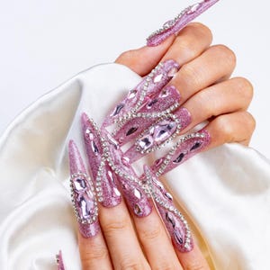 May include: Long, glittery pink artificial nails with large, sparkling rhinestones and gems. The nails are a stiletto shape and are decorated with various sizes of clear and pink crystals. The nails are on a white satin background.