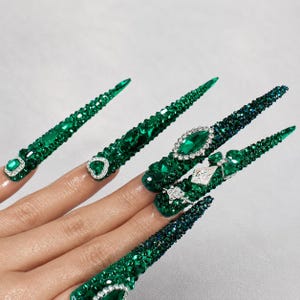 May include: Long, stiletto-shaped press-on nails featuring a deep emerald green color. The surface is heavily embellished with various sizes of green rhinestones, crystals, and silver-toned accents, creating a highly textured, sparkling appearance.