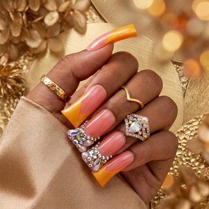 May include: A hand with long, square-shaped nails painted in a peach and yellow gradient design. The nails are adorned with rhinestones and pearls. Gold rings, including a V-shaped ring and a square ring with a large stone, are also visible.