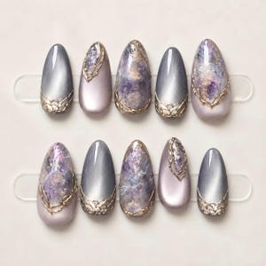May include: A set of ten almond-shaped press-on nails featuring a mix of solid metallic grey, soft lavender, and marbled purple and white designs. Several nails are accented with intricate, raised gold filigree detailing along the edges.