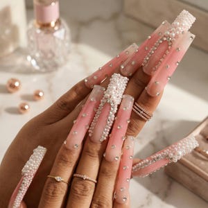 May include: Long, pink and white ombre acrylic nails with pearl and rhinestone embellishments. The nails are adorned with various sizes of pearls and rhinestones, creating a glamorous and elegant look. Several rings are also visible.