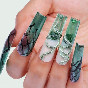 May include: Long, square-shaped artificial nails featuring a translucent green finish with black web-like patterns. Two nails display a three-dimensional silver snake coiled around the surface, accented with green glitter and a white base gradient.