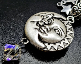 May include: A silver-toned pendant featuring a detailed sun and moon design, with a crystal cube charm hanging below. The sun and moon are depicted with facial features. The pendant is attached to a chain with star-shaped charms.