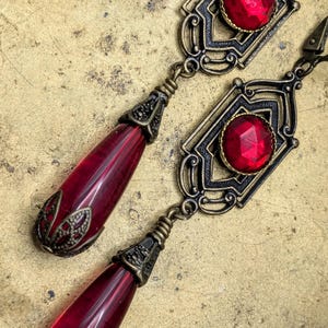 May include: A pair of ornate earrings featuring a geometric design with a large, faceted red gemstone at the center. The earrings have a teardrop-shaped red glass pendant. The metalwork is dark with bronze accents.