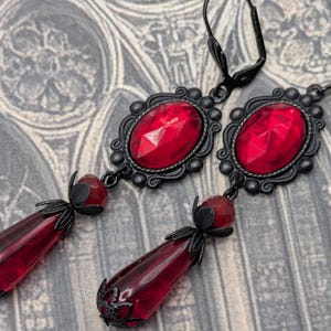 May include: A pair of gothic-style earrings with a dark, ornate design. Each earring features a large, faceted red gem set in a black filigree frame. Below the gem hangs a teardrop-shaped red glass pendant, adding a dramatic flair.