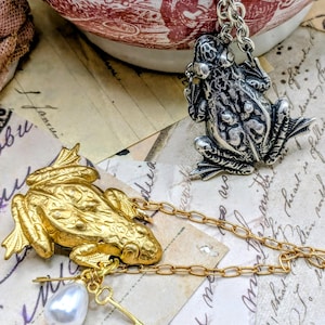 May include: Two frog pendants. One is gold-toned with a pearl and key charm, the other is silver-toned. Both are on chains. The background includes vintage paper and a red and white patterned bowl.