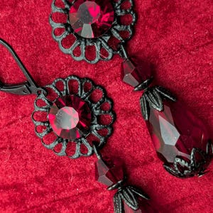 May include: A pair of ornate drop earrings with a dark red gemstone and black filigree detailing. The earrings feature a large, faceted teardrop-shaped stone suspended from a floral-shaped stud. The background is a textured red fabric.