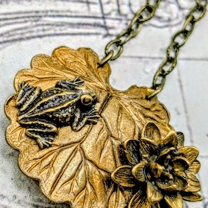 May include: A brass-toned pendant necklace featuring a frog on a lily pad with a flower. The frog is detailed with black accents. The lily pad and flower have intricate textures. The chain is a matching brass color.