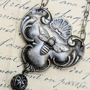 May include: Silver-toned necklace featuring a detailed bee pendant with a shell accent. The pendant is attached to a chain and has a decorative, openwork bead. The necklace is set against a vintage paper background.