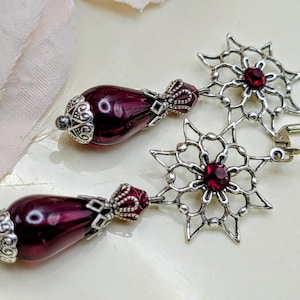 May include: A pair of dangle earrings featuring a dark purple teardrop-shaped bead with ornate silver-colored caps and accents. The earrings also have a silver-colored starburst design with a red gemstone center and a lever-back closure.