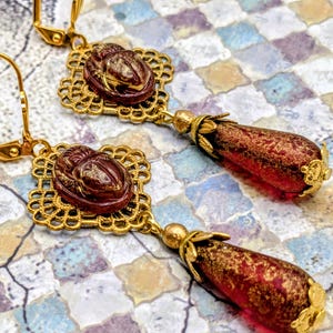 May include: Gold-toned earrings with a scarab beetle design. The earrings feature a square filigree setting with a carved scarab beetle in a reddish-brown hue. Teardrop-shaped red and gold beads dangle below, creating an elegant, vintage aesthetic.
