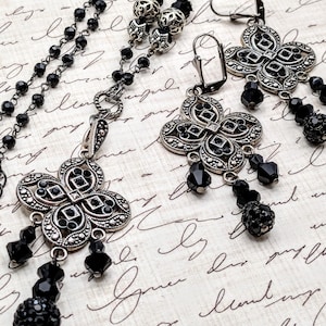 May include: A jewelry set featuring a necklace and earrings. The necklace and earrings have a silver-tone metalwork design with black accents and dangling black beads. The earrings have a lever-back closure. The jewelry is displayed on a cream-colored background.