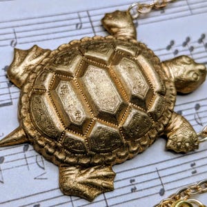 May include: A gold-colored turtle pendant necklace. The turtle pendant has a detailed shell design and is attached to a delicate gold chain. The necklace is displayed on a background of sheet music.