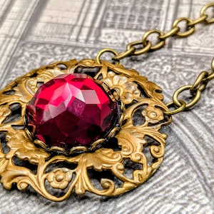 May include: A vintage-style necklace featuring a large, faceted, ruby-red gemstone pendant. The pendant is set in an ornate, gold-toned filigree frame with floral and scroll details. The necklace has a matching gold-toned chain.