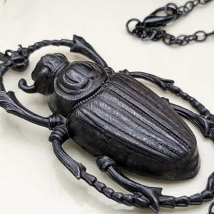 May include: A black beetle pendant necklace. The pendant is a detailed, three-dimensional insect with textured wings and legs. The necklace chain is also black and has a simple link design. The necklace is likely made of metal.