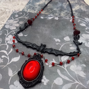 May include: A black gothic necklace featuring a bat pendant and a large red oval-shaped stone. The necklace has red bead accents and a decorative frame around the stone. The necklace is designed with a dark, vintage aesthetic.