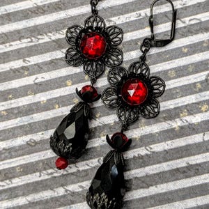 May include: A pair of dangle earrings featuring black metal filigree flower settings with central faceted red crystals. Below each flower hangs a large, faceted black teardrop bead, accented with small red beads at the top and bottom.