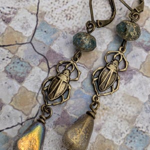 May include: A pair of antique brass-toned earrings. Each earring features a beetle-shaped charm, a blue-green bead, and a teardrop-shaped bead. The teardrop beads have a metallic, iridescent finish. The earrings have a lever-back closure.