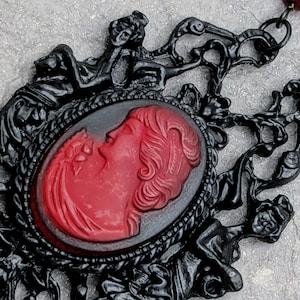 May include: A black filigree pendant featuring a central oval cameo with a red profile portrait of a person. The pendant is attached to a chain with dark red beads, showcasing intricate scrollwork and detailed relief carvings.