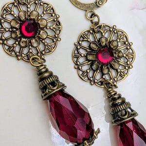 May include: A pair of antique-style earrings. Each earring features a filigree flower design with a red gemstone center and a teardrop-shaped, faceted, deep red crystal pendant. The earrings have a bronze finish and lever-back closures.