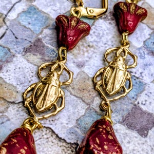 May include: Gold-toned earrings with a unique design. Each earring features a red, flower-shaped top, a gold scarab beetle charm, and a red, teardrop-shaped bead. The earrings are suspended from gold-colored ear wires.