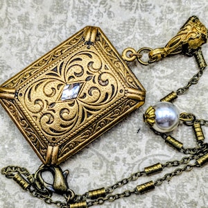May include: An antique gold-toned rectangular pendant necklace with intricate scrollwork and a central diamond-shaped gem. The necklace features a delicate chain with a pearl accent and a decorative clasp, showcasing vintage charm.