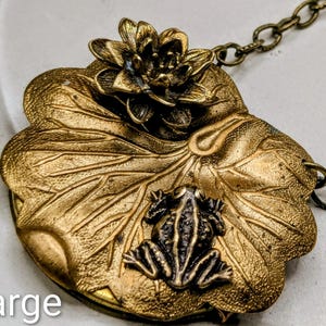 May include: A gold-toned metal pendant necklace featuring a detailed lily pad design. A small frog sits on the lily pad, with a flower at the top. The word "large" is at the bottom. The chain is a matching antique brass color.