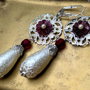 May include: A pair of ornate dangle earrings. Each earring features a silver filigree flower with a burgundy center and a pearl accent. Below, a burgundy bead connects to a pearlescent, teardrop-shaped dangle.