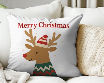 May include: White decorative pillow with a cartoon reindeer design. The reindeer has brown antlers, a red nose, and wears a red and white striped hat and a green sweater. The words "Merry Christmas" are printed in red at the top.