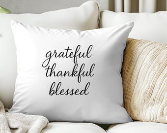May include: White square pillow with the words "grateful, thankful, blessed" in black script. The pillow is on a white sofa with a beige throw blanket and a second pillow.
