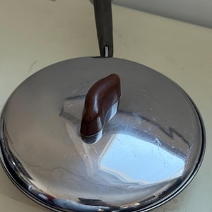 Puede incluir: Una tapa de acero inoxidable con un asa marrón oscuro y un pomo marrón más pequeño. La tapa es redonda y tiene una superficie brillante y reflectante. El asa está sujeta a la tapa con un lazo metálico.