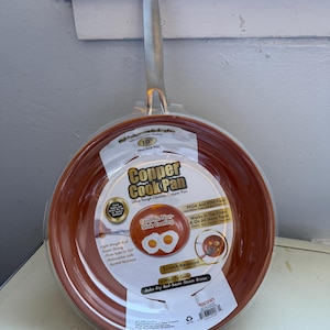 Könnte beinhalten: Eine kupferfarbene 25 cm Antihaft-Bratpfanne mit silbernem Griff. Die Pfanne hat eine keramische Antihaftbeschichtung und ist kratzfest. Die Verpackung hebt die Eigenschaften der Pfanne hervor, einschließlich der Ofen- und Spülmaschinenfestigkeit.