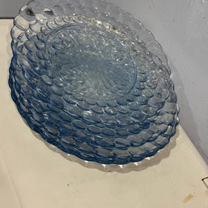 Puede incluir: Una pila de platos de vidrio azul claro. Cada plato tiene un patrón texturizado en forma de burbujas alrededor del borde y un diseño central. Los platos están apilados, mostrando sus intrincados detalles y color.
