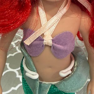 May include: A doll with red hair and a purple top. The doll's top is made of a purple felt material and is tied with a ribbon. The doll has a green sequined skirt and a white collar.