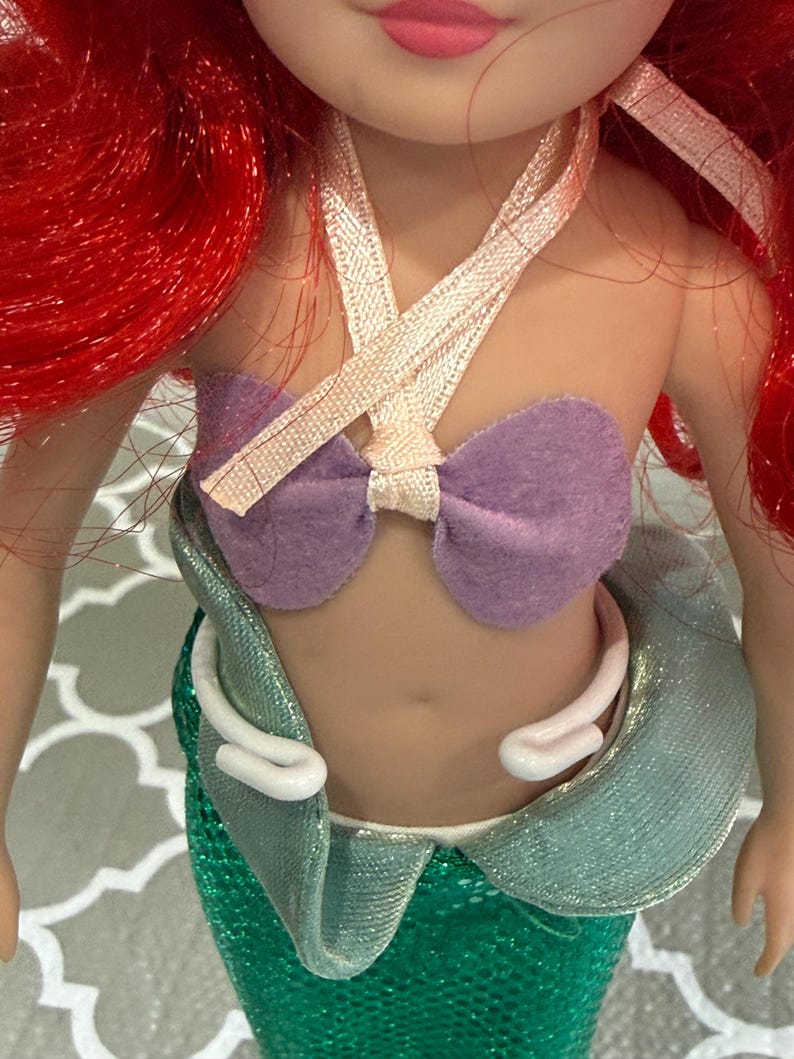May include: A doll with red hair and a purple top. The doll's top is made of a purple felt material and is tied with a ribbon. The doll has a green sequined skirt and a white collar.