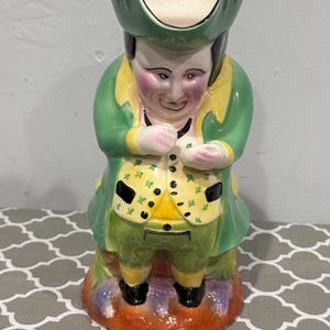 Puede incluir: Jarra de cerámica con forma de hombre que lleva un abrigo verde, un chaleco amarillo y zapatos negros. La jarra tiene un interior blanco y un sombrero verde. El hombre sostiene un pequeño objeto verde, posiblemente un trébol. La base es una mezcla de morado y naranja.