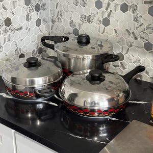 Puede incluir: Un juego de tres piezas de cocina de acero inoxidable que incluye una olla a presión alta y dos ollas más bajas, todas con tapas negras a juego con pomos y una banda decorativa de círculos geométricos en colores rojo, naranja y negro.
