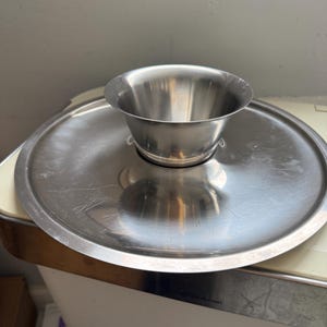May include: A stainless steel serving set featuring a round tray with a raised, centered bowl. The tray and bowl have a brushed metal finish. The bowl is designed to sit securely in the center of the tray. Ideal for serving snacks or dips.