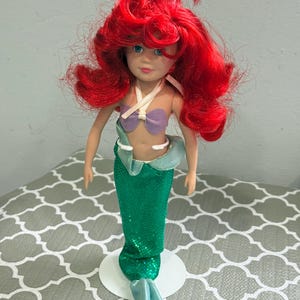May include: A doll of a mermaid with bright red hair, blue eyes, and a purple seashell top. The doll has a green sequined tail and is standing on a white base. The mermaid doll is a popular children's toy.