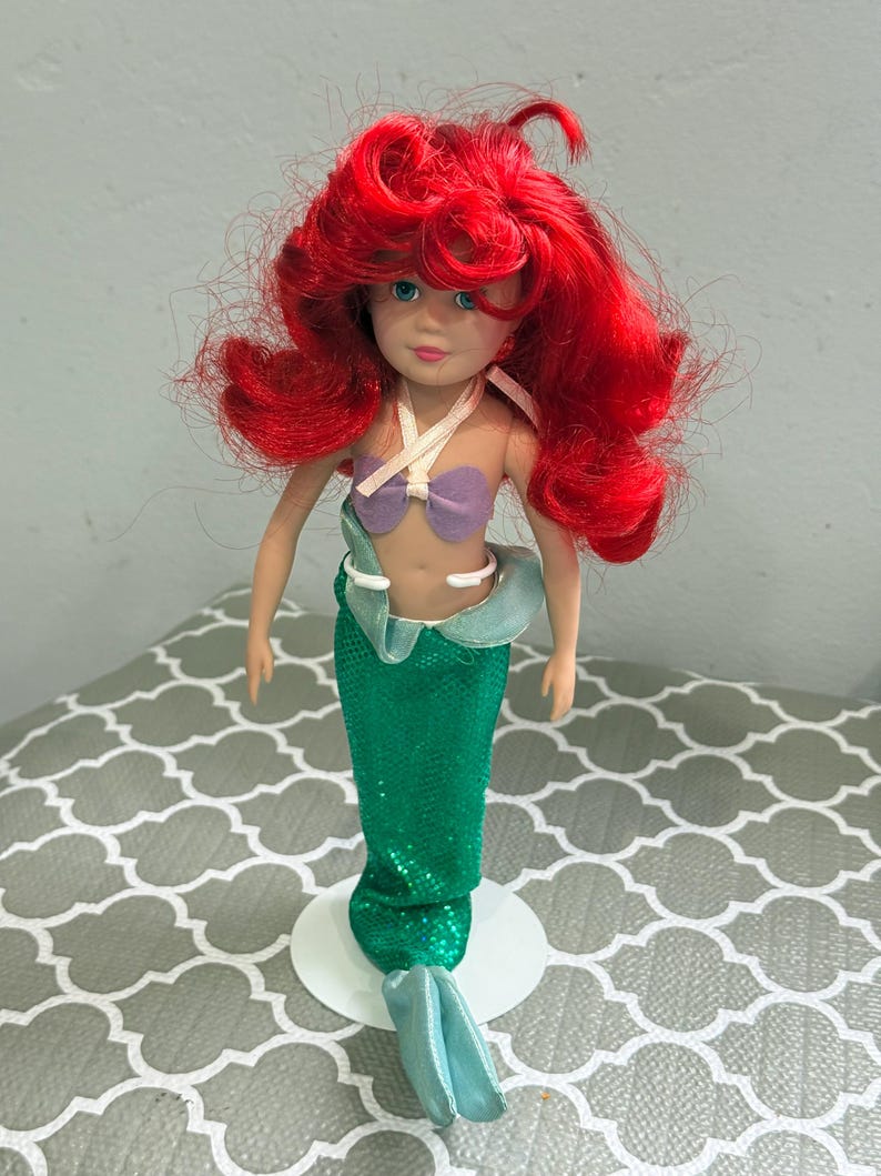 May include: A doll of a mermaid with bright red hair, blue eyes, and a purple seashell top. The doll has a green sequined tail and is standing on a white base. The mermaid doll is a popular children's toy.
