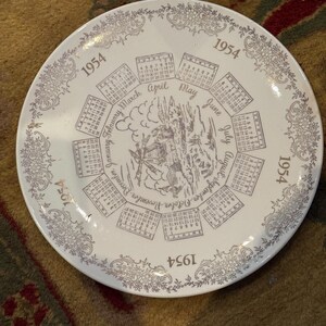 Könnte beinhalten: Weiße Keramikplatte mit silbernem Blumenrand und dem Jahr 1954, das sich am Rand wiederholt. Die Mitte zeigt ein Kalenderdesign mit Monaten und eine zentrale Illustration eines Gebäudes und Bäume.