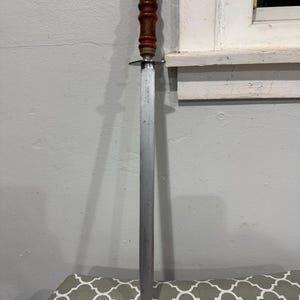 K&ouml;nnte beinhalten: Ein Vintage-Stahl-Messersch&auml;rfer mit Holzgriff und Messingakzenten. Der Griff ist dunkelbraun und die Klinge ist lang und schlank. Der Sch&auml;rfer dient zum Sch&auml;rfen von Messern.