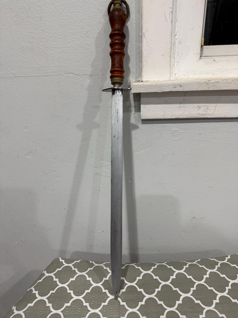 K&ouml;nnte beinhalten: Ein Vintage-Stahl-Messersch&auml;rfer mit Holzgriff und Messingakzenten. Der Griff ist dunkelbraun und die Klinge ist lang und schlank. Der Sch&auml;rfer dient zum Sch&auml;rfen von Messern.