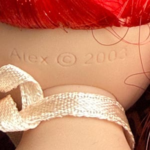 May include: A doll's neck and head with red hair. The doll's neck is adorned with a cream-colored ribbon. The doll's head has the name "Alex" and the copyright symbol and the year "2003" imprinted on the back of the neck.