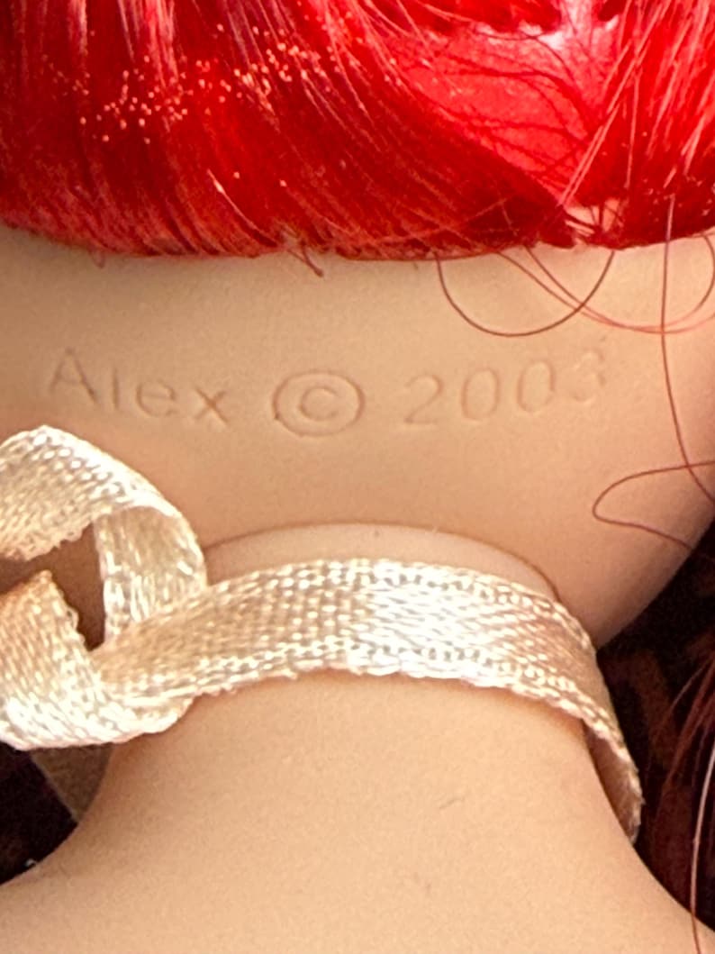 May include: A doll's neck and head with red hair. The doll's neck is adorned with a cream-colored ribbon. The doll's head has the name "Alex" and the copyright symbol and the year "2003" imprinted on the back of the neck.